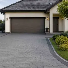Piękny podjazd z kostki brukowej prowadzi do garażu i wejścia do domu. Wokół zadbany ogród z krzewami i drzewem.